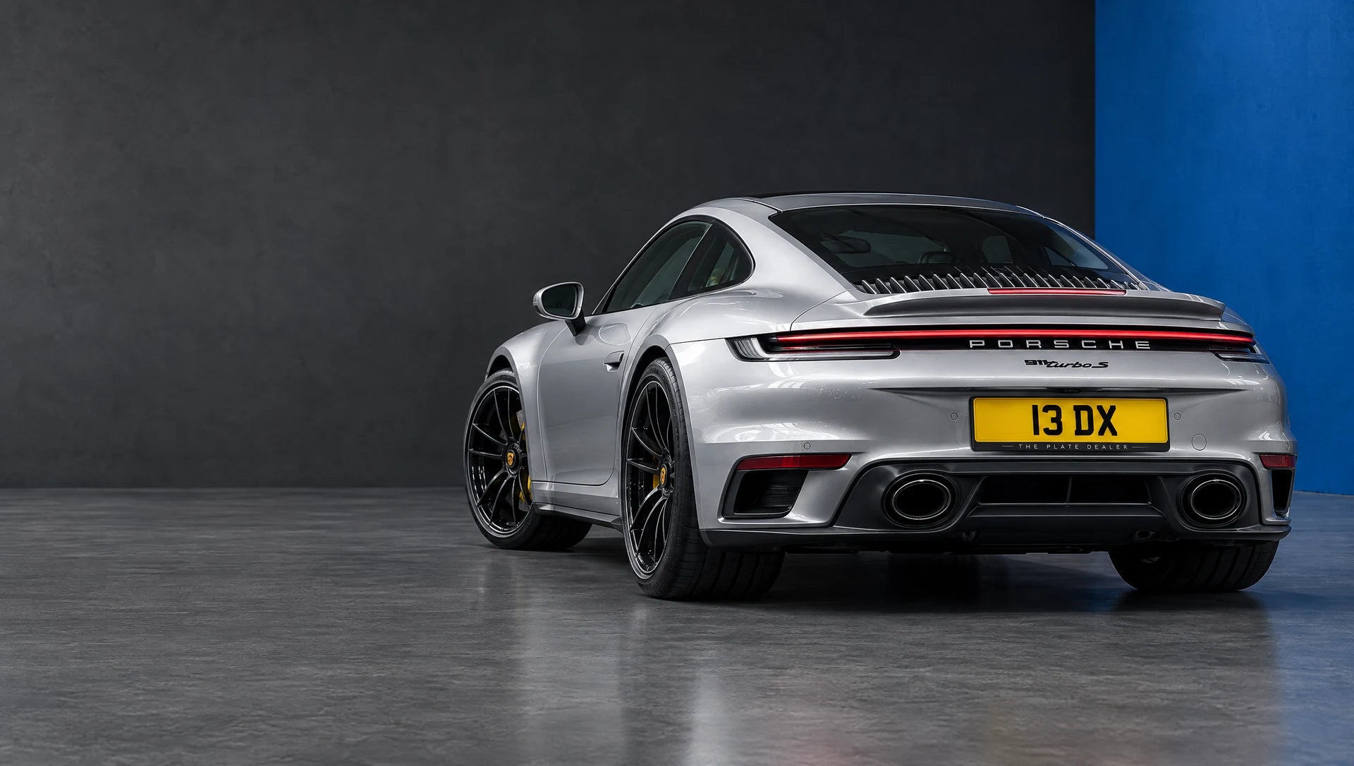 Silver Porsche 911 displaying the private registration 13 DX in The Plate Dealer's blue and charcoal studio